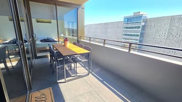 Een balkon met een tafel en stoelen, een glazen deur die naar een kamer leidt, en een uitzicht op gebouwen in de verte.