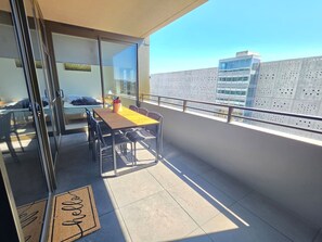 Een balkon met een tafel en stoelen, een glazen deur die naar een kamer leidt, en een uitzicht op gebouwen in de verte.