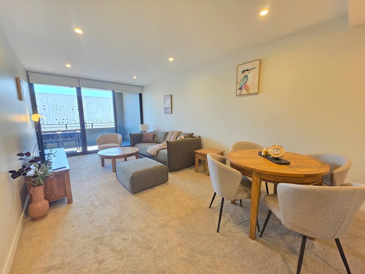 Un salon moderne comprenant un canapé, un pouf, une table basse et une table à manger avec des chaises. On y trouve également une plante en pot et un tableau accroché au mur.