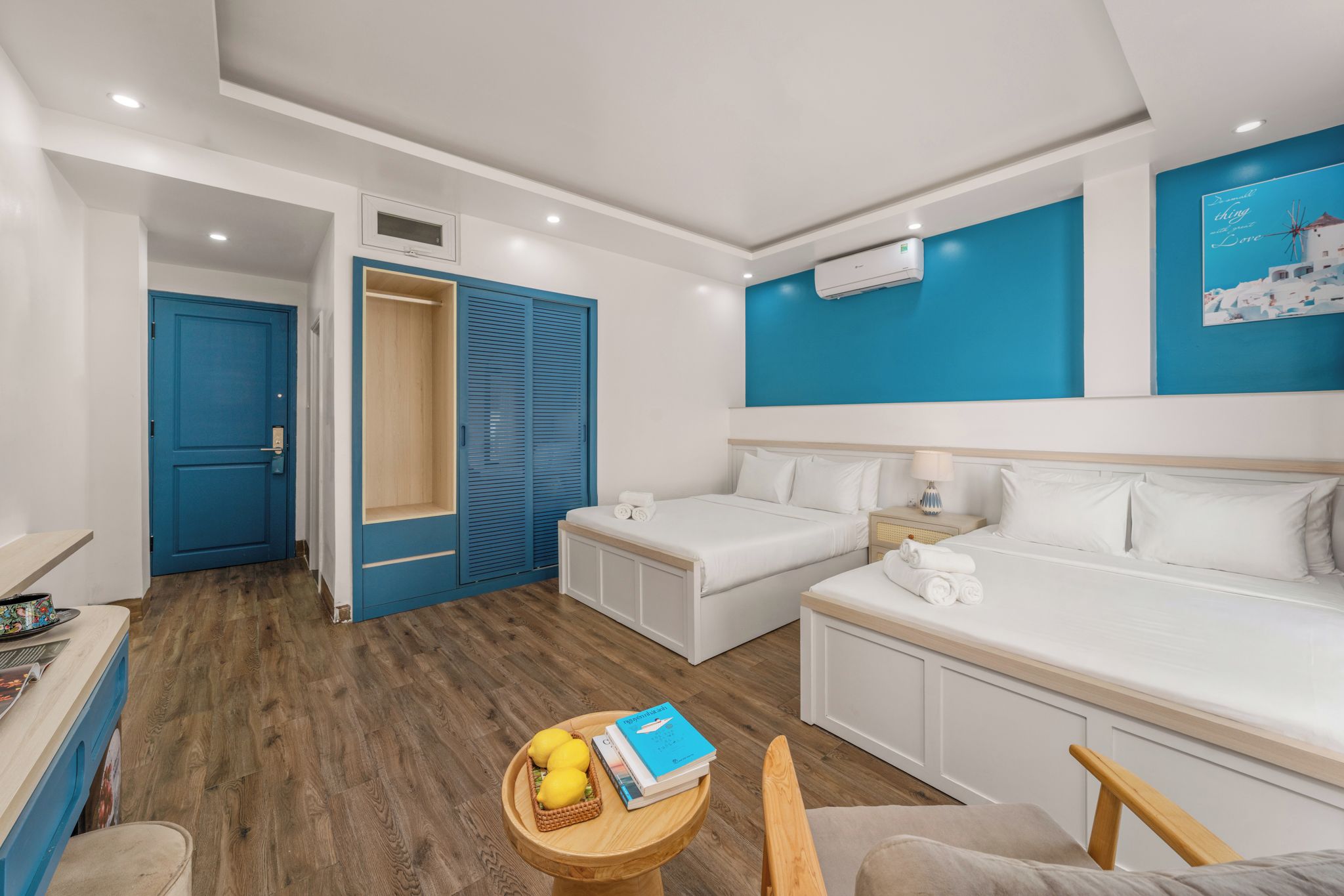 A hotel room with a bed, a desk, a chair, a small table with books and lemons, and a wardrobe.