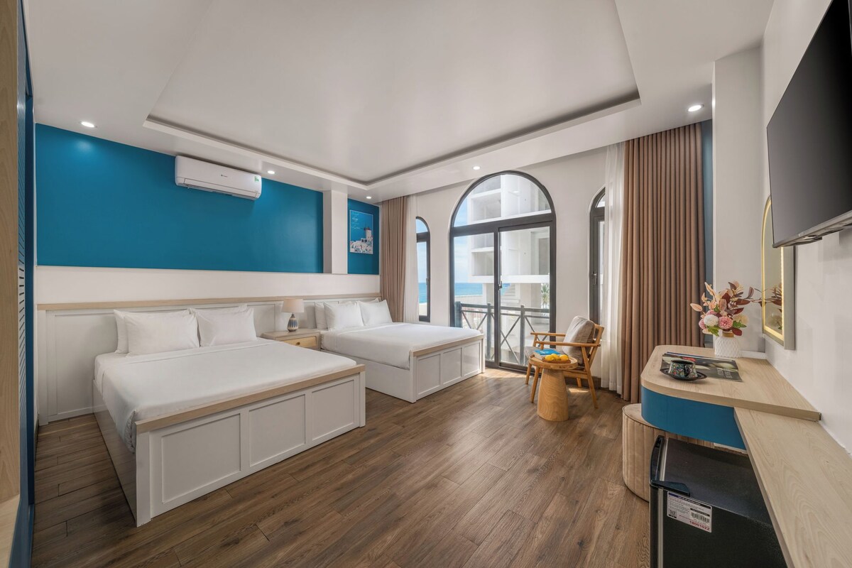 A hotel room with two beds, a small seating area, a TV, and a view of the sea.