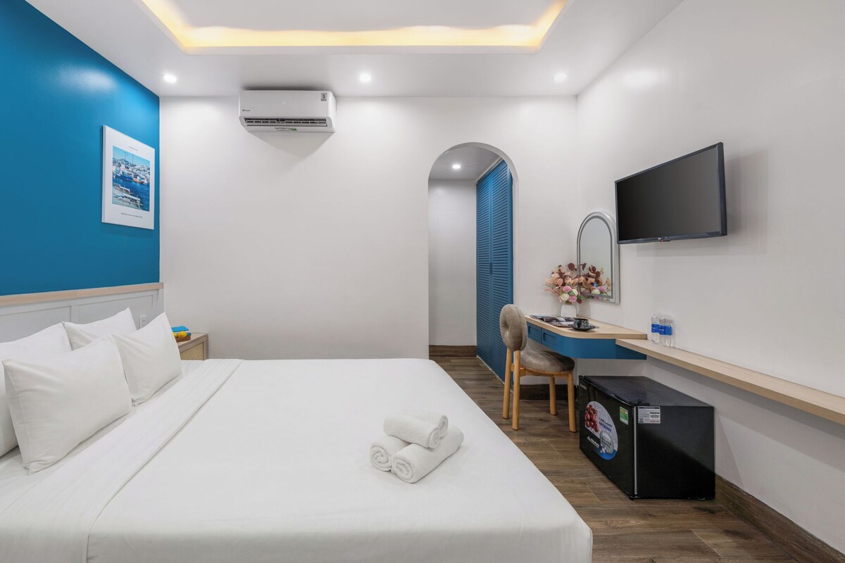 A hotel room with a bed, a desk, a chair, a TV, and an air conditioning unit.