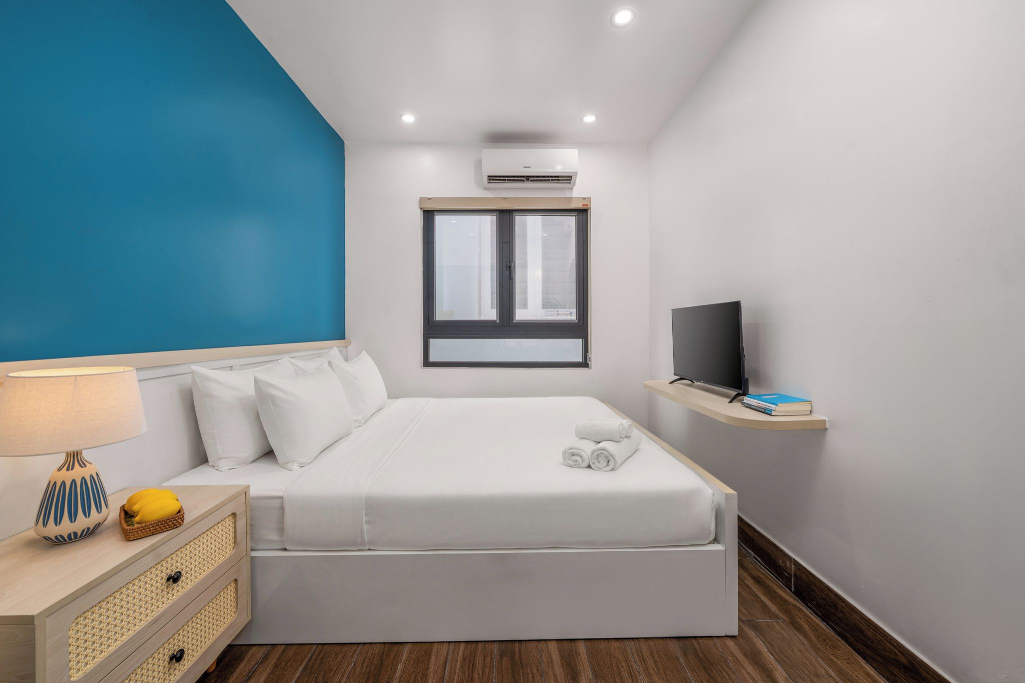 A modern hotel room with a bed, a television on a shelf, a window, and an air conditioning unit.