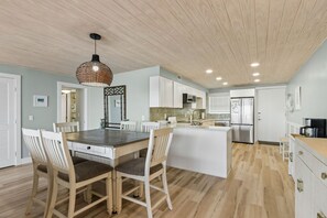A modern kitchen with white cabinetry, a central island, and a dining area with wooden chairs. - Comfy Upper Unit Condo To Enjoy The Beach Or The Fishing 2 Bedroom Condo (Fernandina Beach)