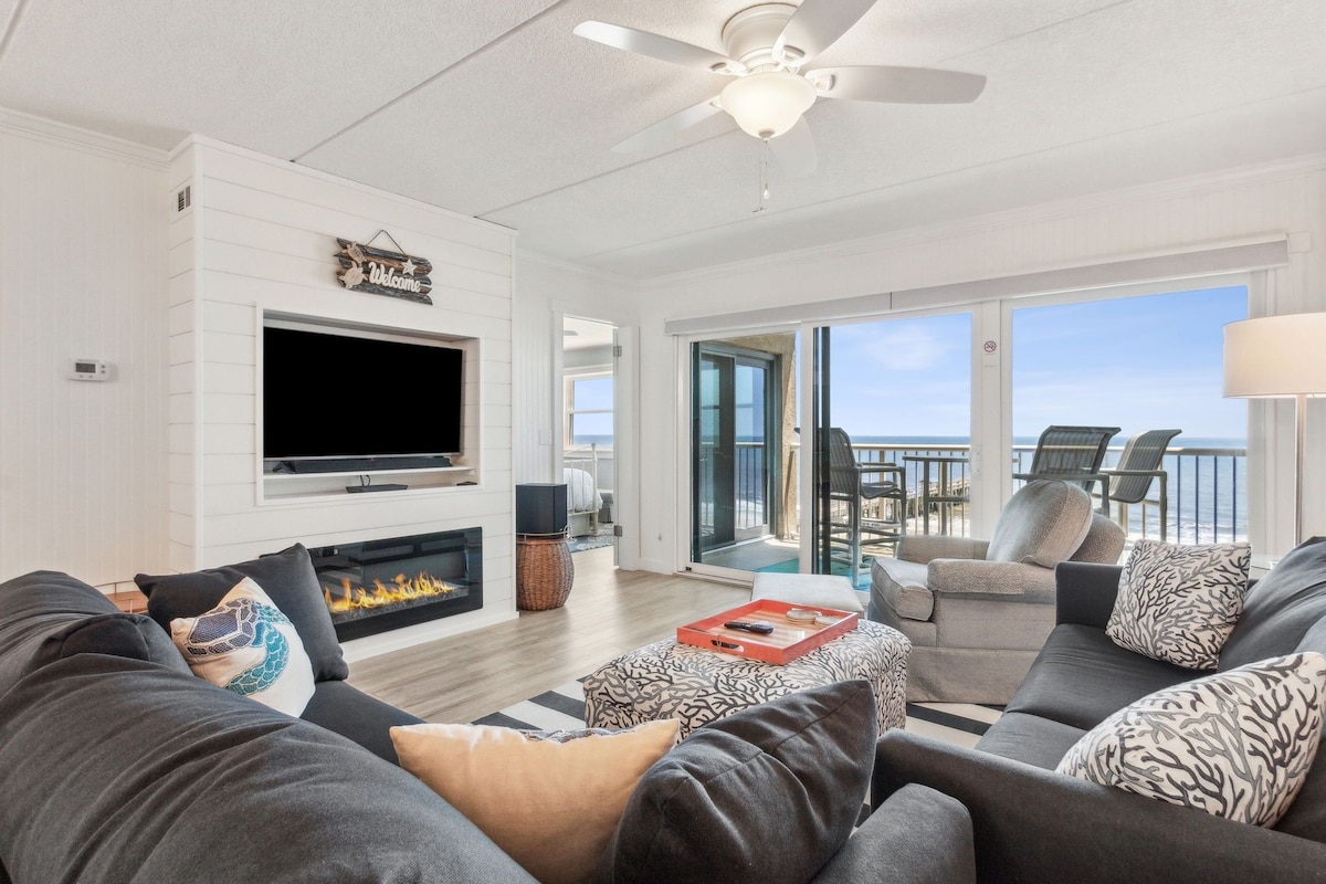 A spacious living room with a flat-screen TV, a fireplace, and a balcony with outdoor seating.
