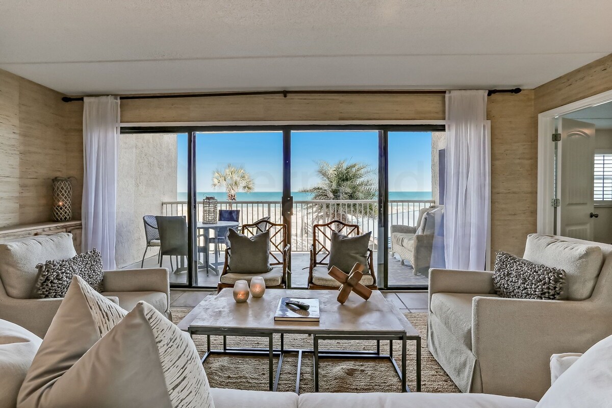 A room with a view of the beach, furnished with a sofa, chairs, and a table.
