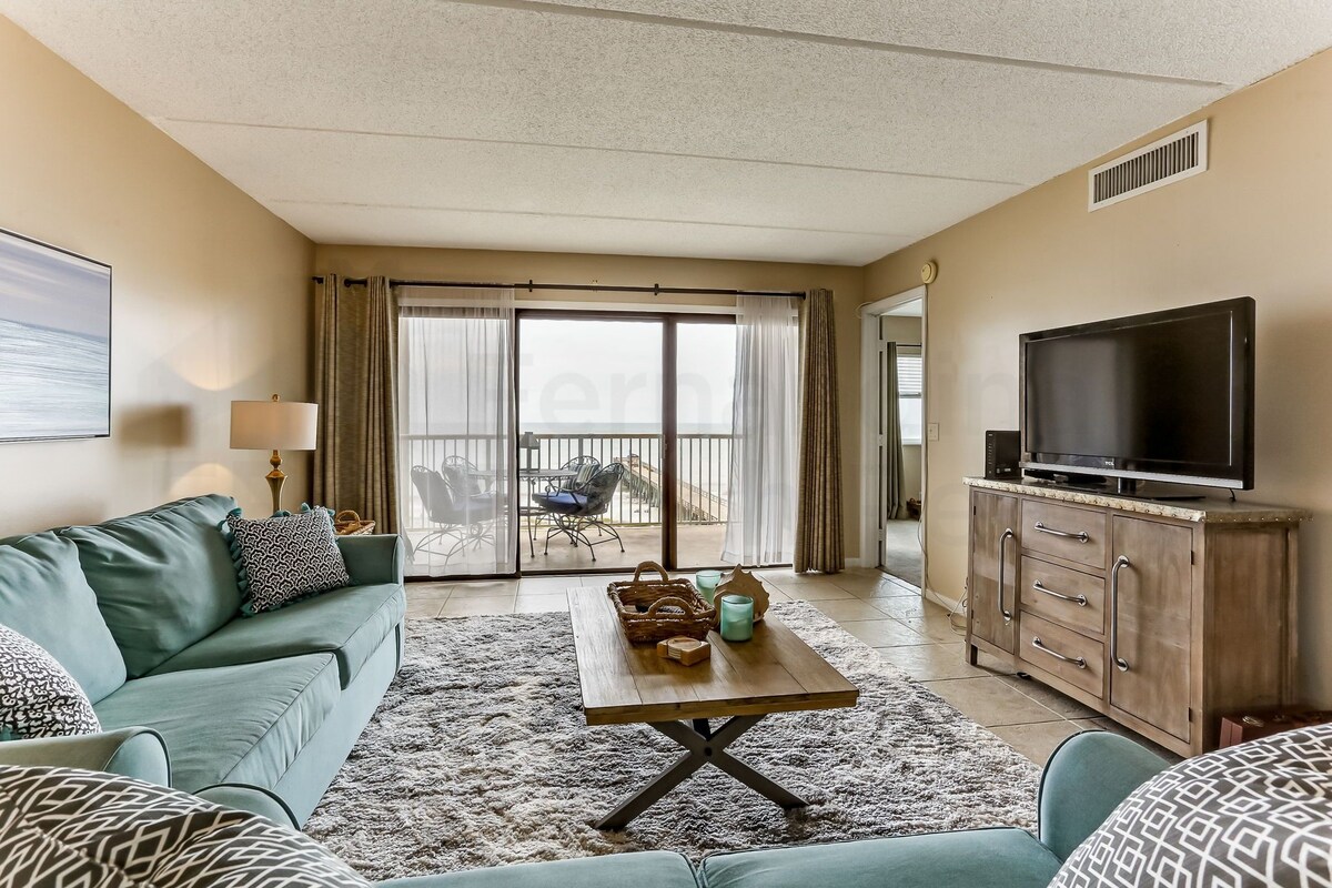 A living room with a teal sofa, a wooden coffee table, a television on a wooden cabinet, and a view through a sliding glass door to an outdoor area.