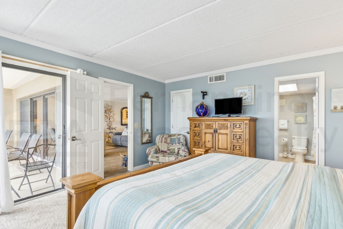 A bedroom with a bed, wooden dresser, television, and a door leading to a balcony.
