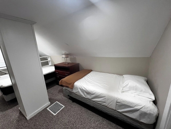 A small, single-bed room with a white mattress, a brown blanket, a bedside table with a lamp, and a window with blinds. - Niagara Riverside Villa Sleeps 8 Scenic Retreat (Niagara Falls)