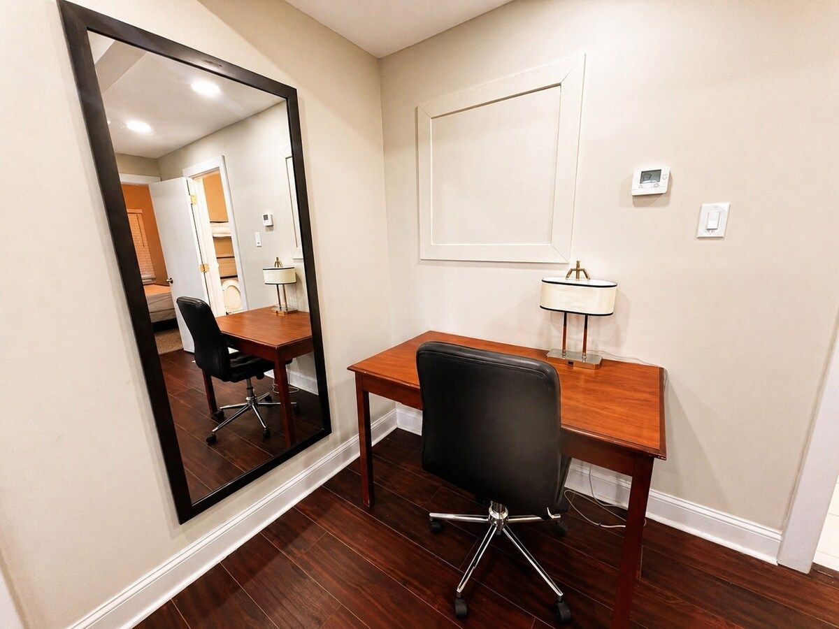 Quarto com escrivaninha de madeira, cadeira de escritório preta, espelho grande e luminária fixa na parede.