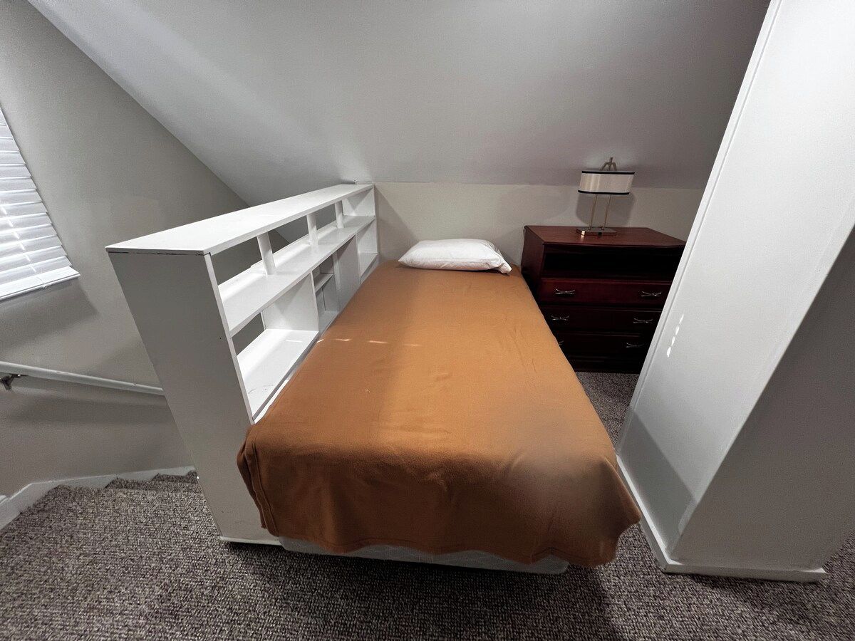 Une petite chambre avec un lit simple, dotée d’une tête de lit blanche, d’une couverture de lit marron, d’un oreiller blanc, d’une table de chevet et d’une lampe.