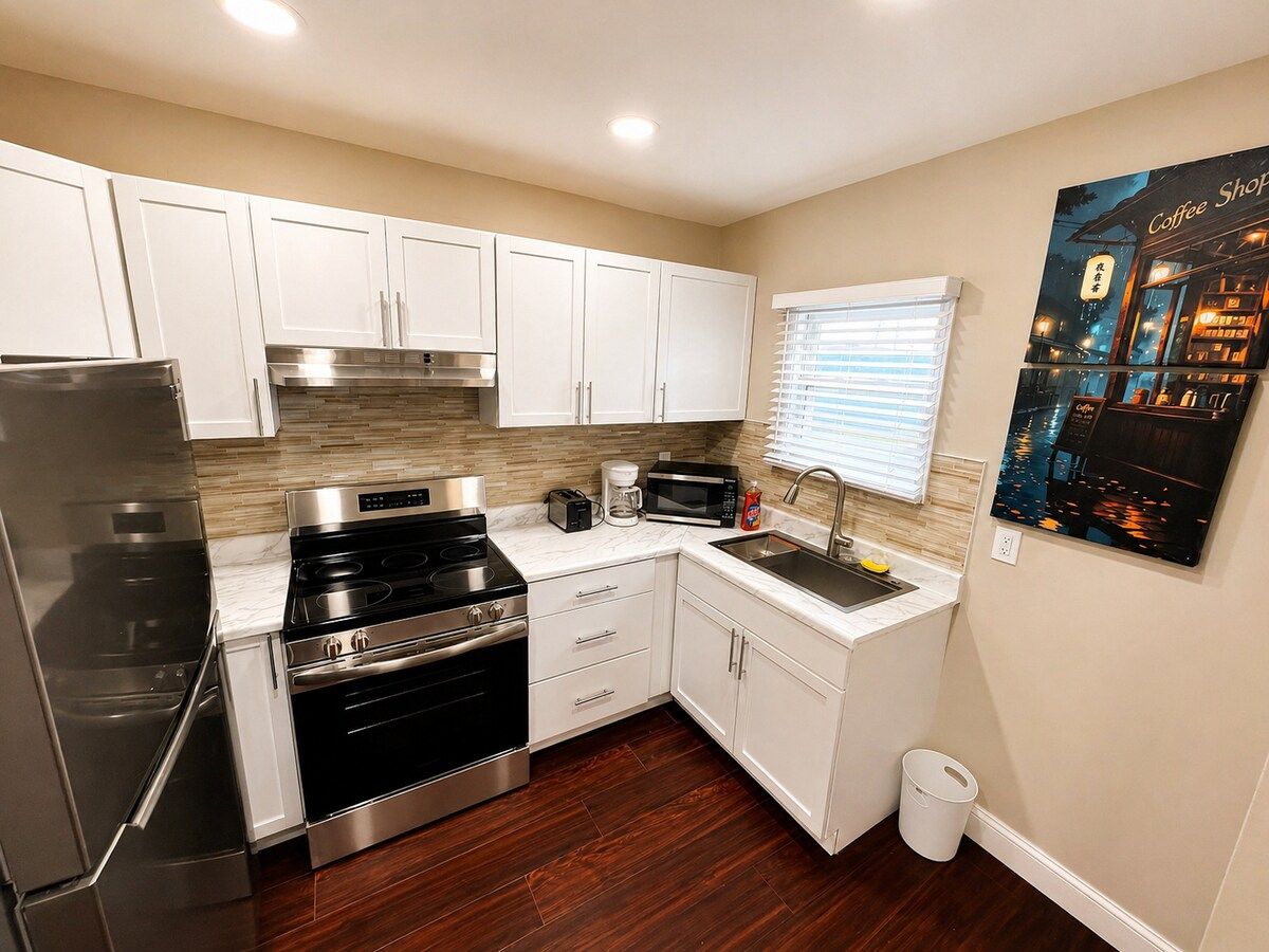 A compact kitchen with white cabinets, stainless steel appliances, and a small window with blinds.