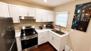A compact kitchen with white cabinets, stainless steel appliances, and a small window with blinds.