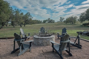 A fire pit with four Adirondack chairs arranged around it, set on a gravel surface with a grassy field and trees in the background.