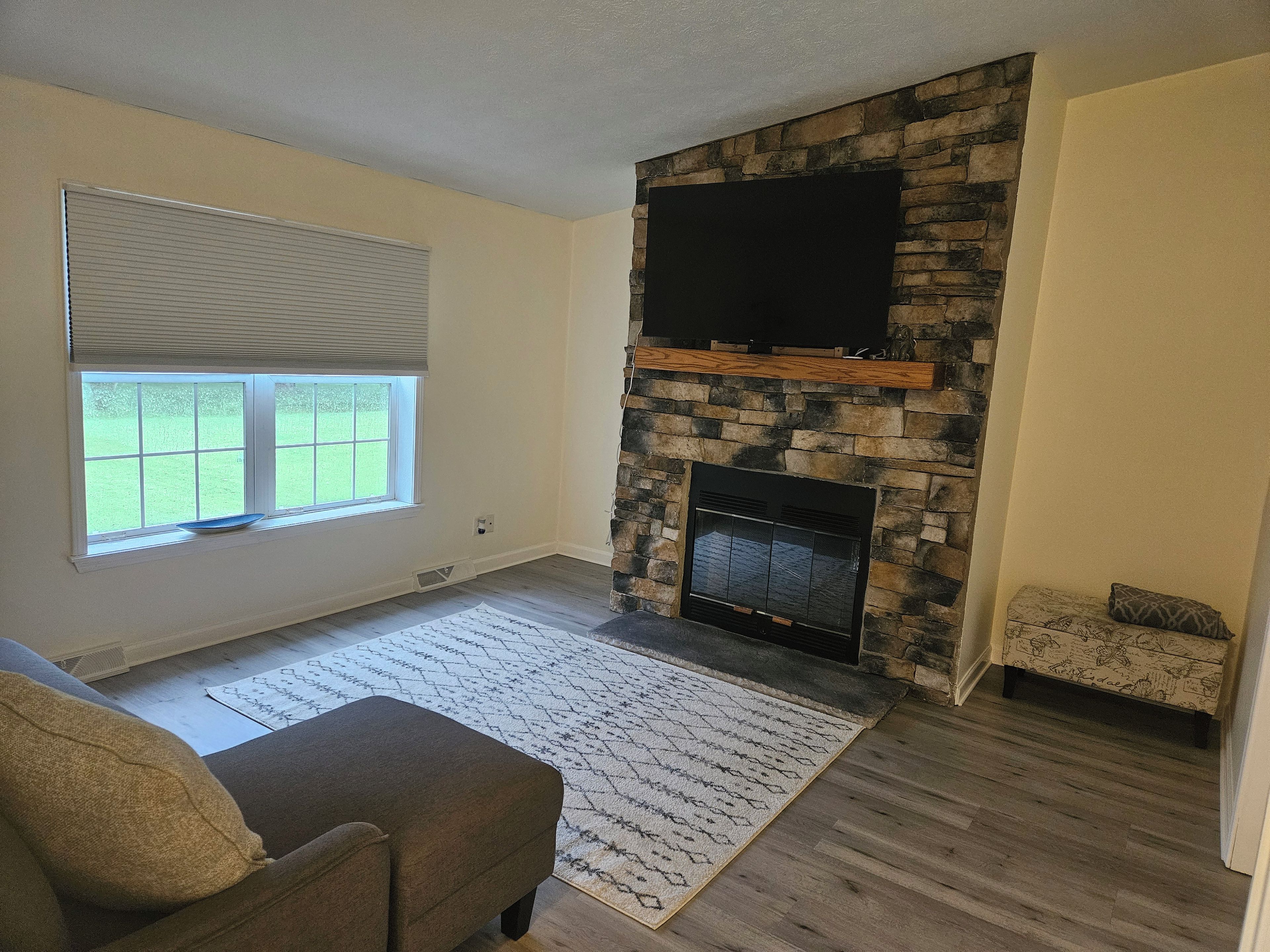 A living room with a stone fireplace, a flat-screen TV mounted above it, a window with blinds, and a patterned rug.