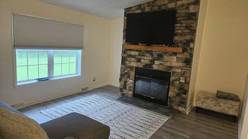 A living room with a stone fireplace, a flat-screen TV mounted above it, a window with blinds, and a patterned rug.