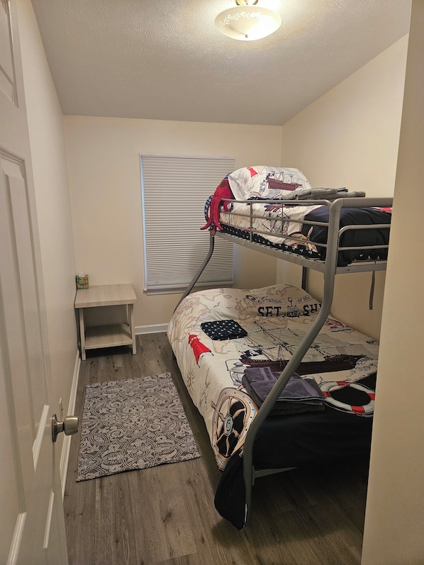 A bunk bed room with a patterned rug, a small table, and a window with blinds.