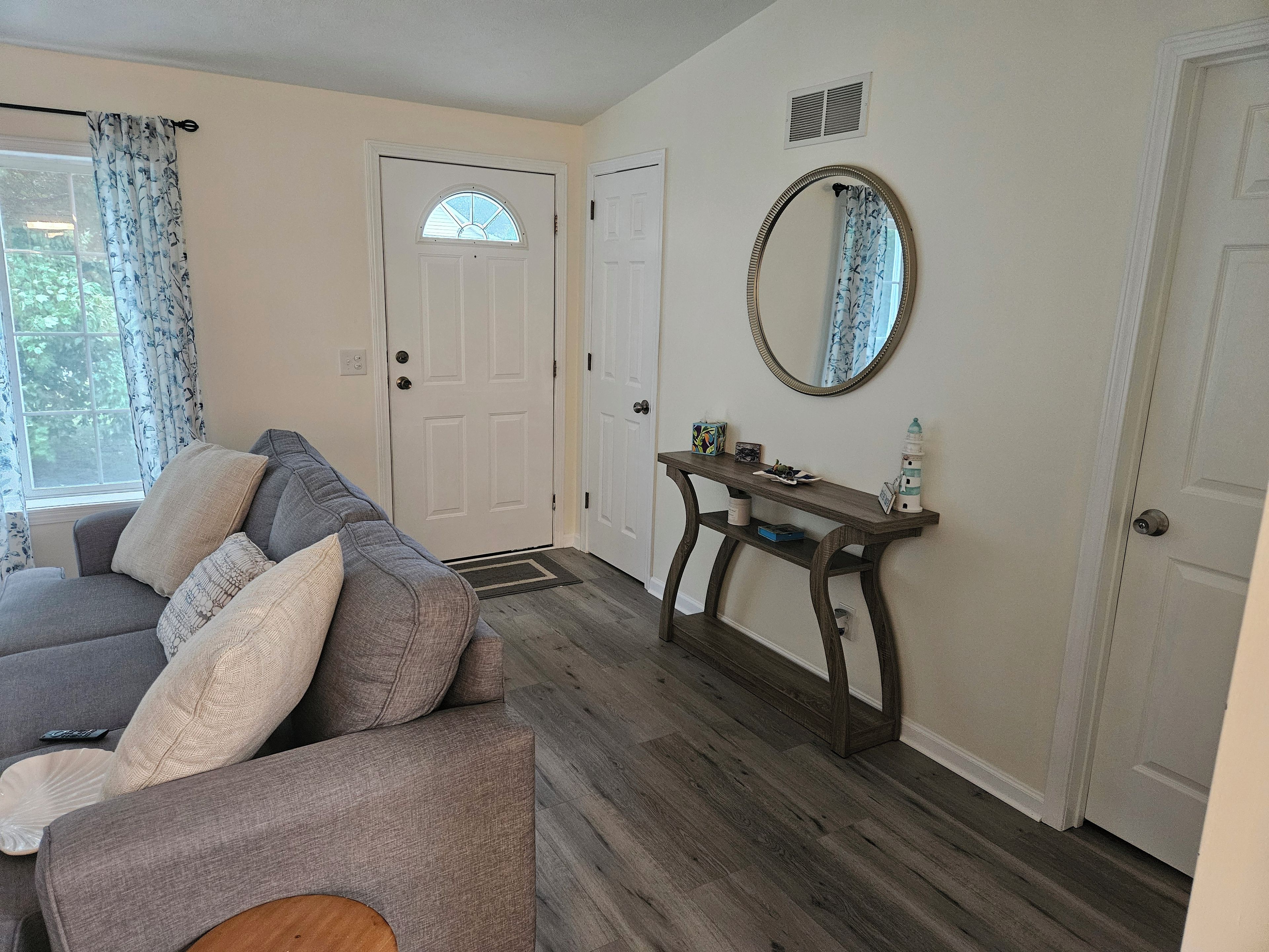 Un salon avec un canapé gris, une console ornée d’objets décoratifs, une porte blanche et une fenêtre avec des rideaux.