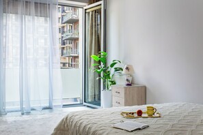 Ein Schlafzimmer mit einem Bett, einem Nachttisch mit Lampe, einem Fenster mit durchsichtigen Vorhängen und Blick auf ein Gebäude.