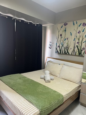 Ein ordentlich bezogenes Bett mit grüner Tagesdecke, einem Teddybären und einem Blumenbild an der Wand.