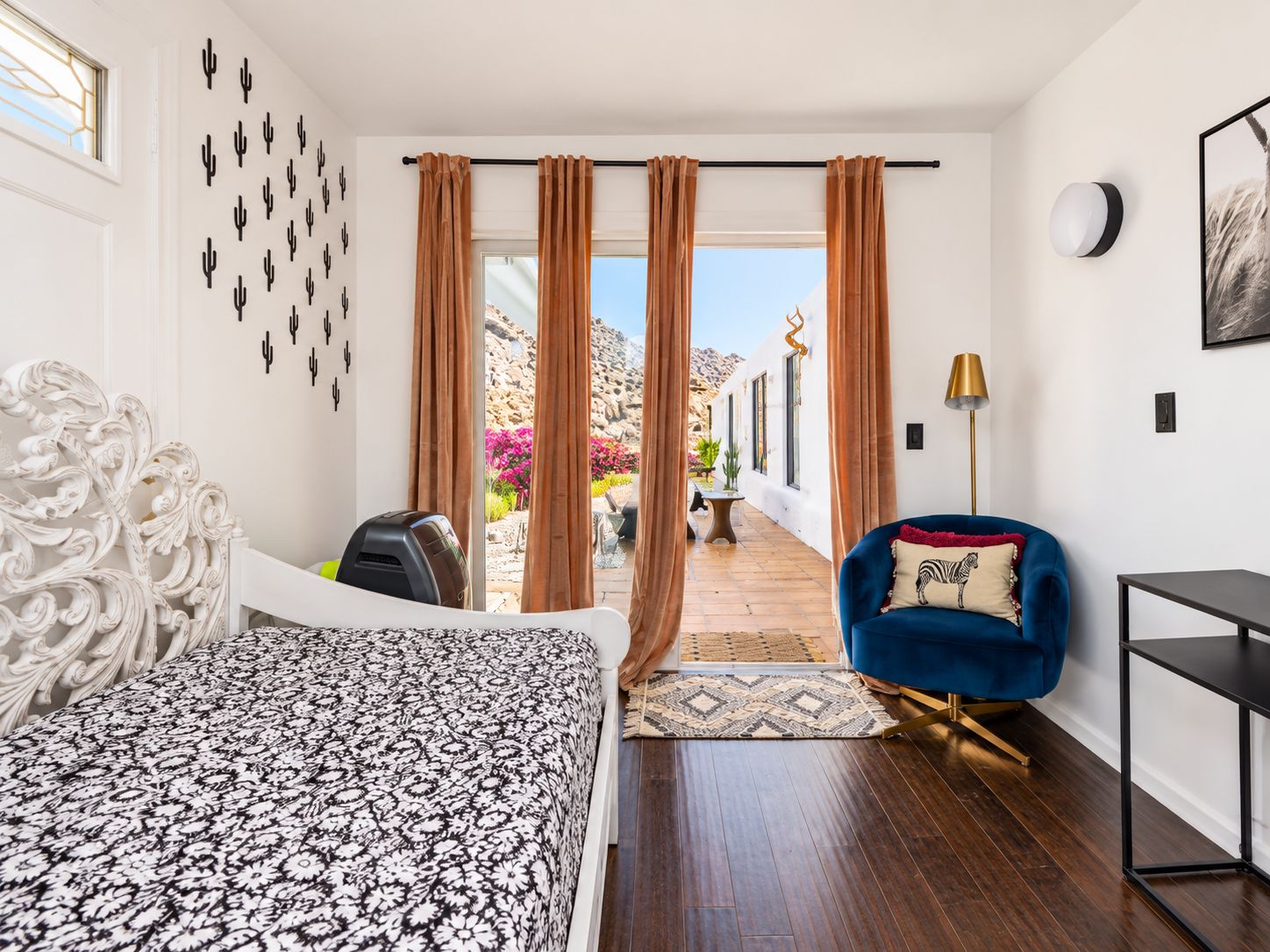A bedroom with a bed, a blue armchair, a desk, and a view of a courtyard with plants and a mountain.
