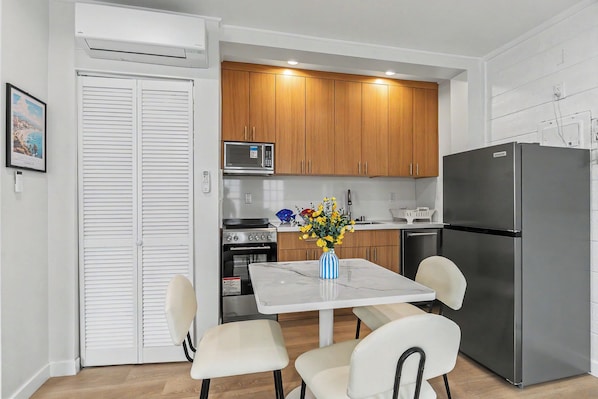 A compact kitchen with wooden cabinets, a stainless steel refrigerator, a microwave, and a stove. There is a dining table with chairs and a vase of flowers.