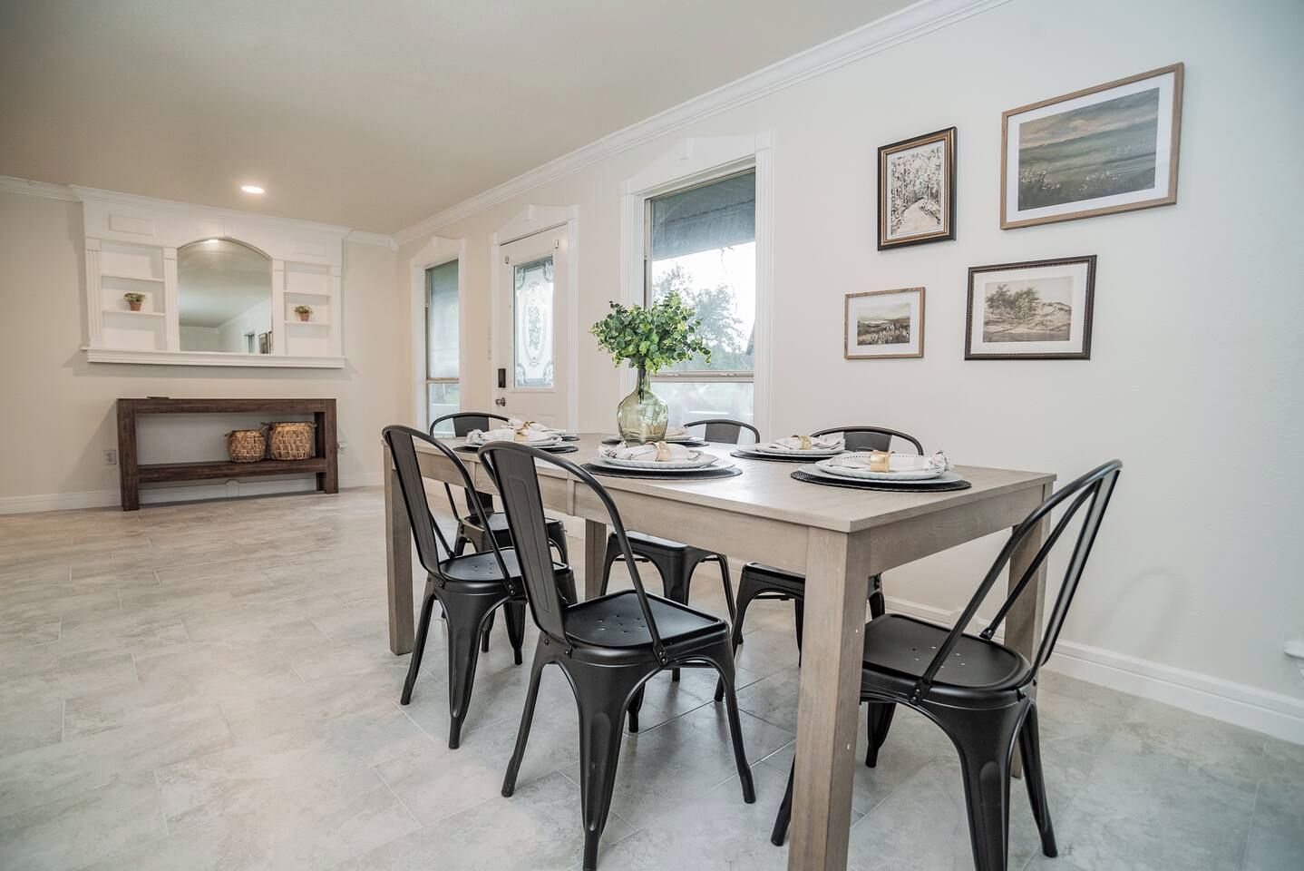 A dining area with a long table, six chairs, and a vase with flowers. There are framed pictures on the wall and a bench with baskets.