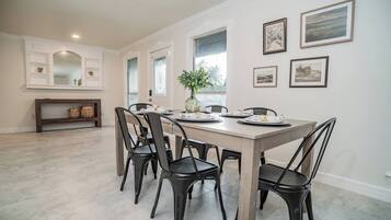 A dining area with a long table, six chairs, and a vase with flowers. There are framed pictures on the wall and a bench with baskets.