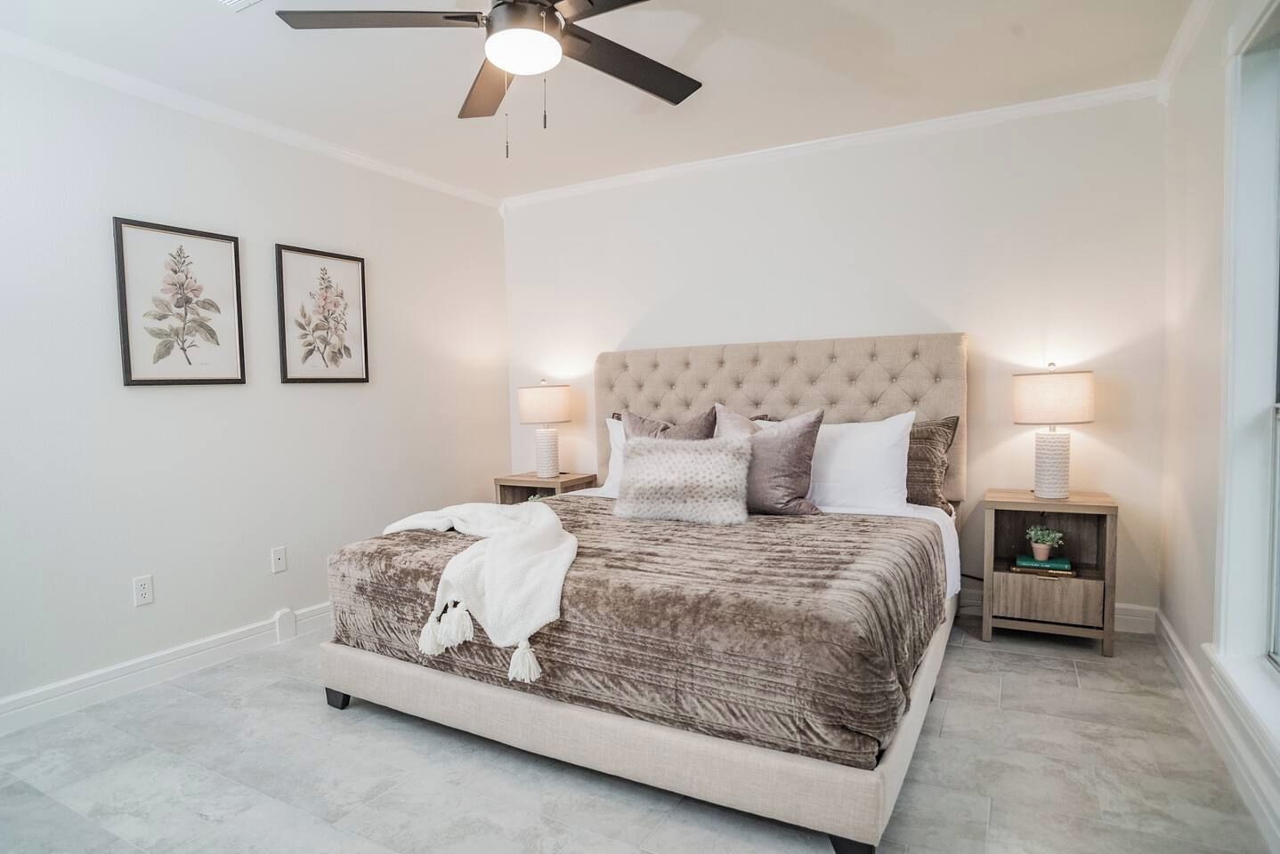 A bedroom with a large bed, two bedside tables with lamps, a ceiling fan, and two framed botanical prints on the wall.