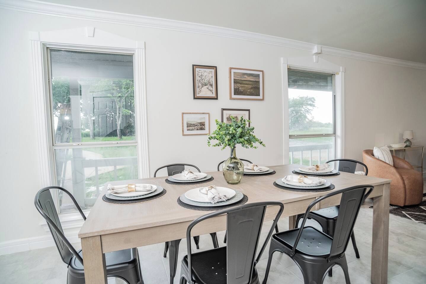 A dining area with a wooden table set for six, surrounded by chairs, with a vase of greenery and framed pictures on the wall.