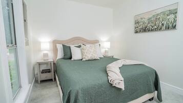 A bedroom with a bed, nightstands, a bedside lamp, a window, and a wall-mounted painting.