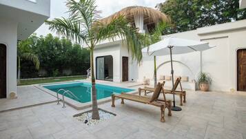 Une piscine avec des chaises longues et un palmier, entourée d’un bâtiment blanc doté d’un toit de chaume.
