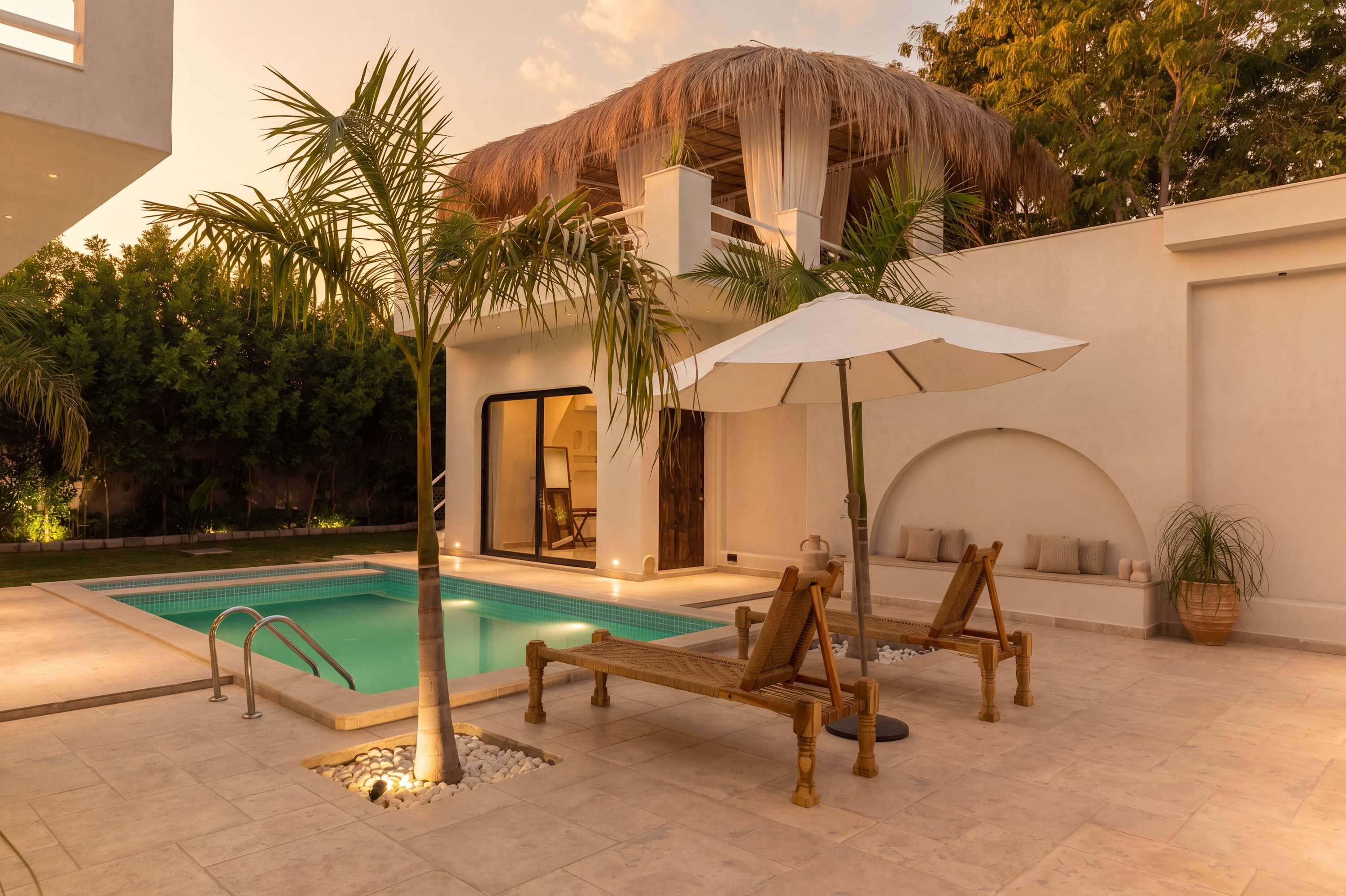 A poolside area with a thatched-roof structure, lounge chairs, and a white umbrella.