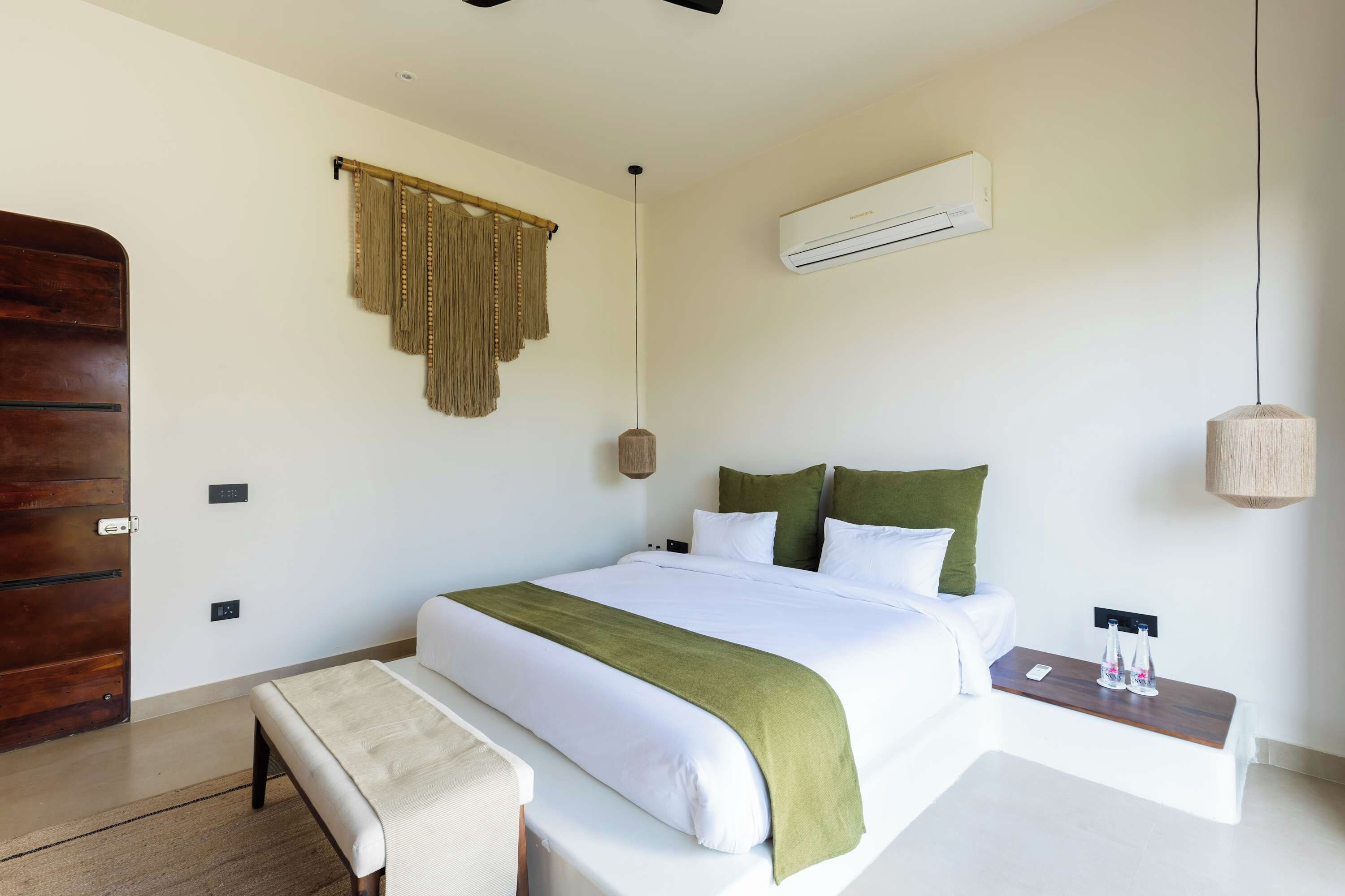 Une chambre à coucher comprenant un lit, une table de chevet, un banc, un climatiseur fixé au mur et une porte en bois.