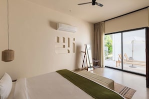 Een hotelkamer met een groot bed, een plafondventilator, een airconditioningapparaat, uitzicht op het buitenterras met een tafel en stoelen, en een groot raam met gordijnen.