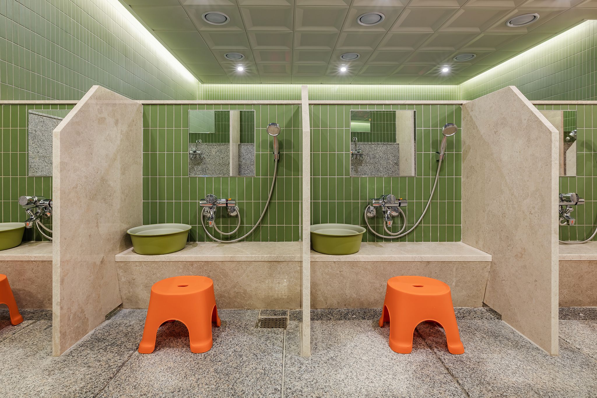A restroom with green tiled walls, individual sinks, and orange stools.