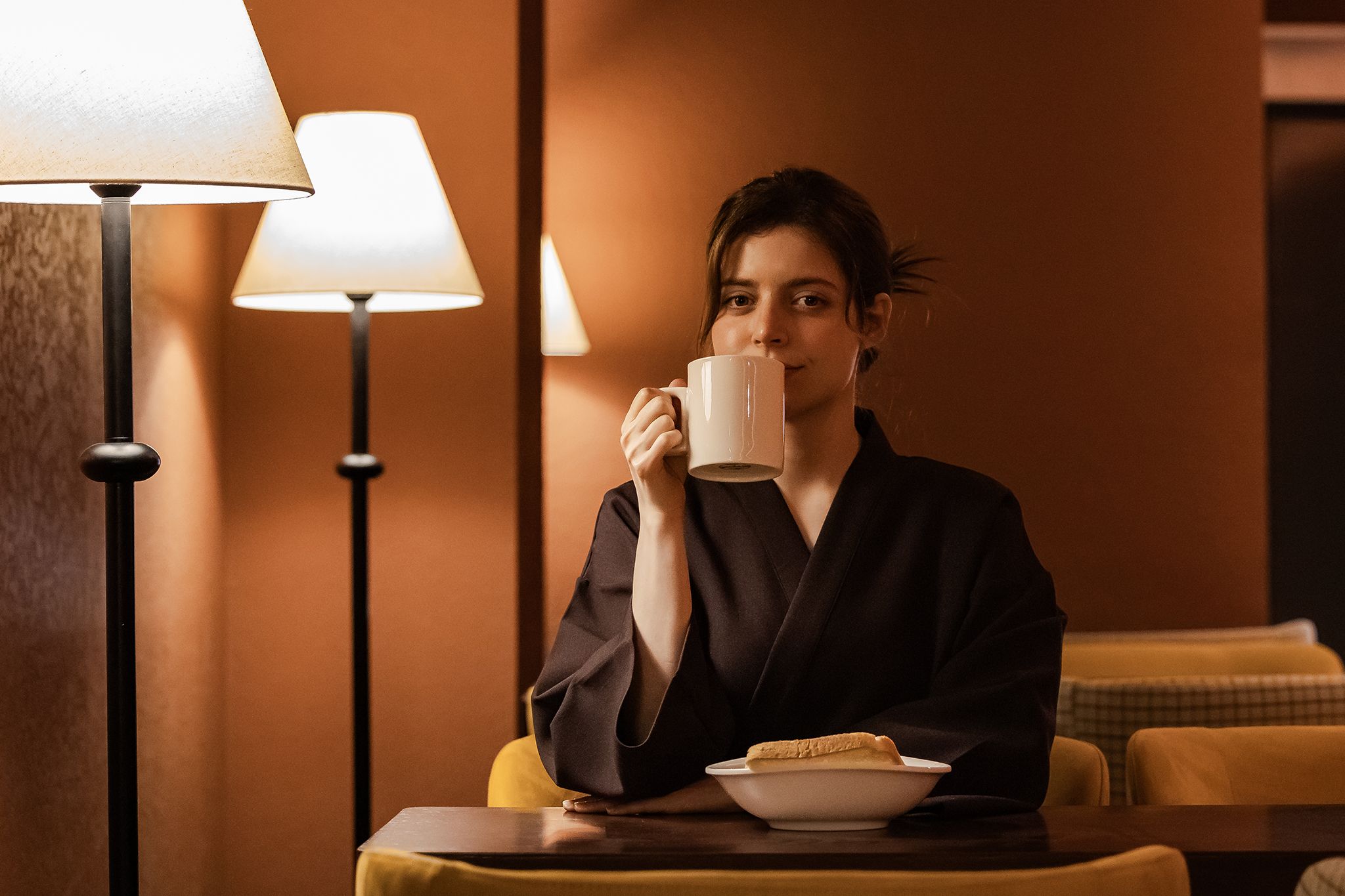 A person sitting at a table with a cup and a plate of food, in a room with a lamp and a sofa.
