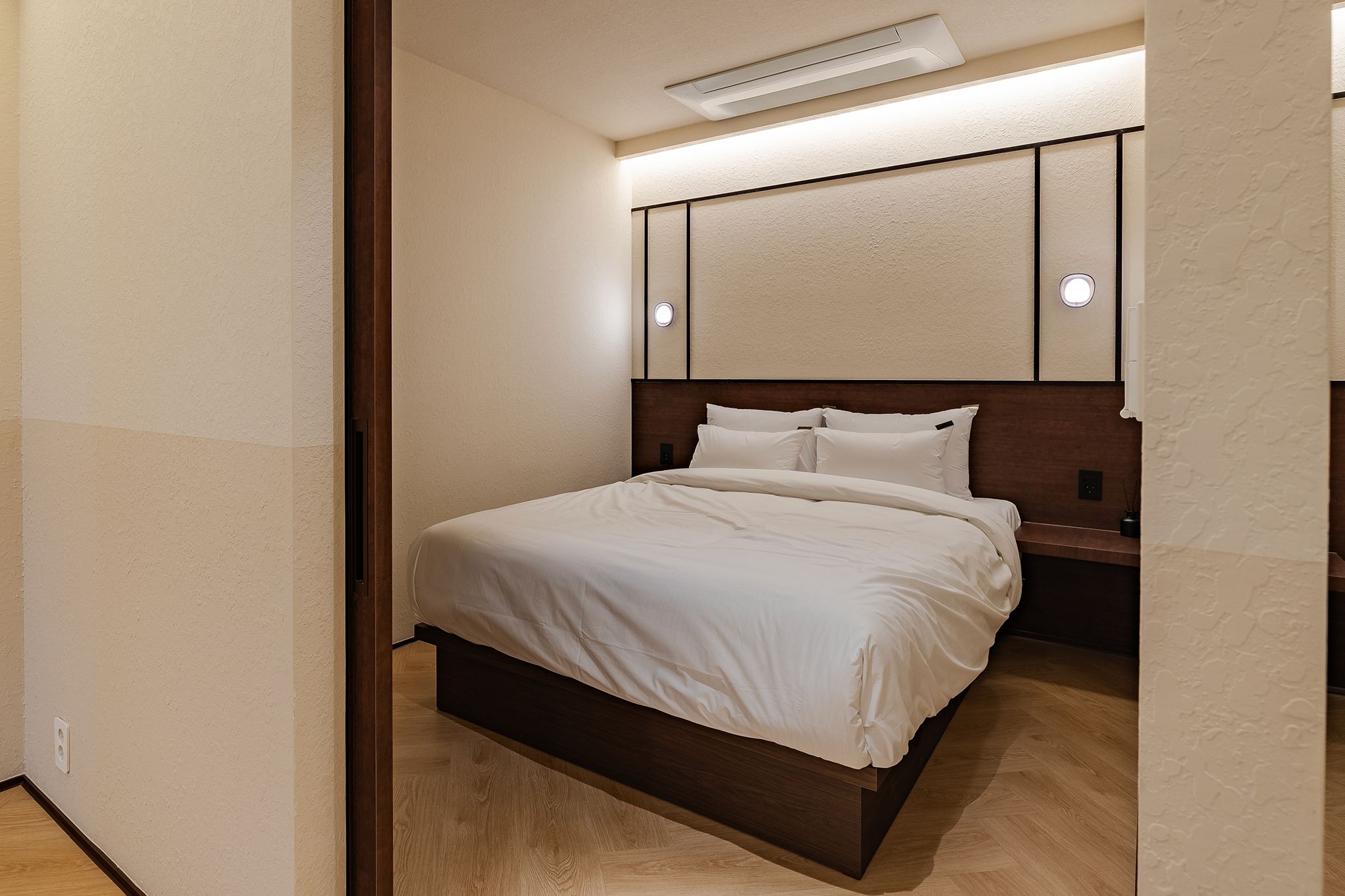 A hotel room with a bed, a nightstand, a wall-mounted light, and a ceiling vent.