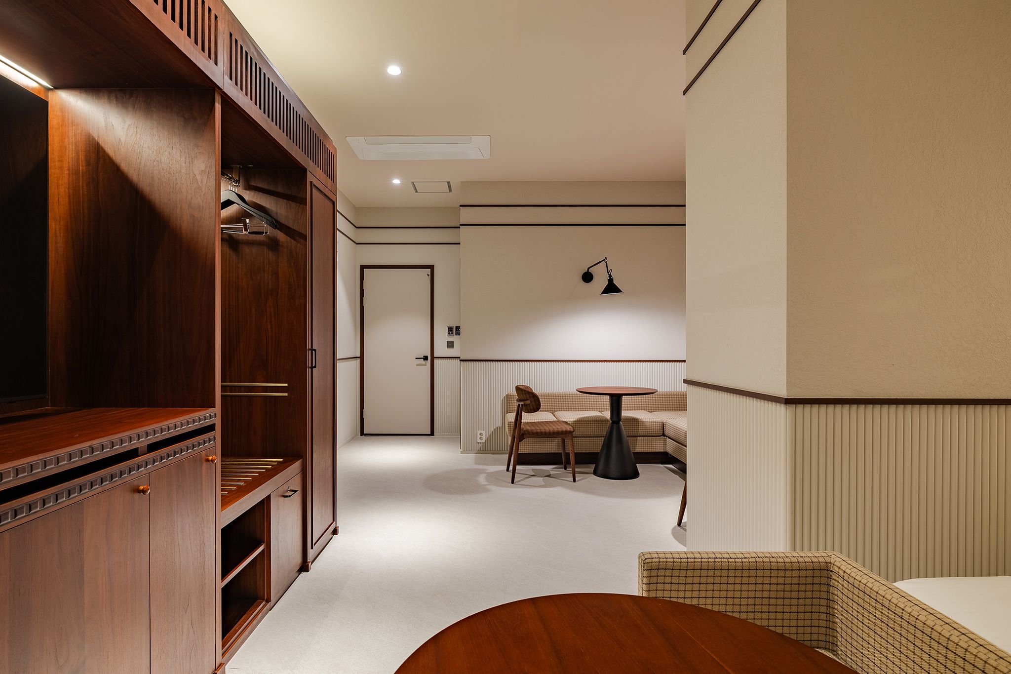 A hotel room with a wooden wardrobe, a round table, a sofa, and a wall-mounted lamp.