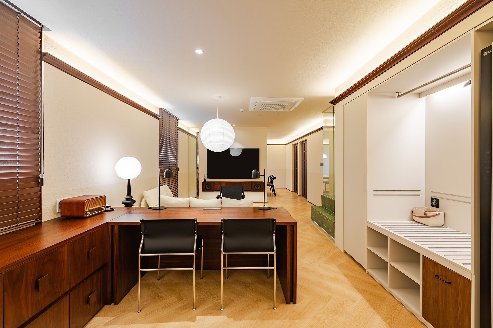 A modern hotel room with a wooden dining table, two chairs, a sofa, a large window with blinds, a flat-screen TV, and a wardrobe.