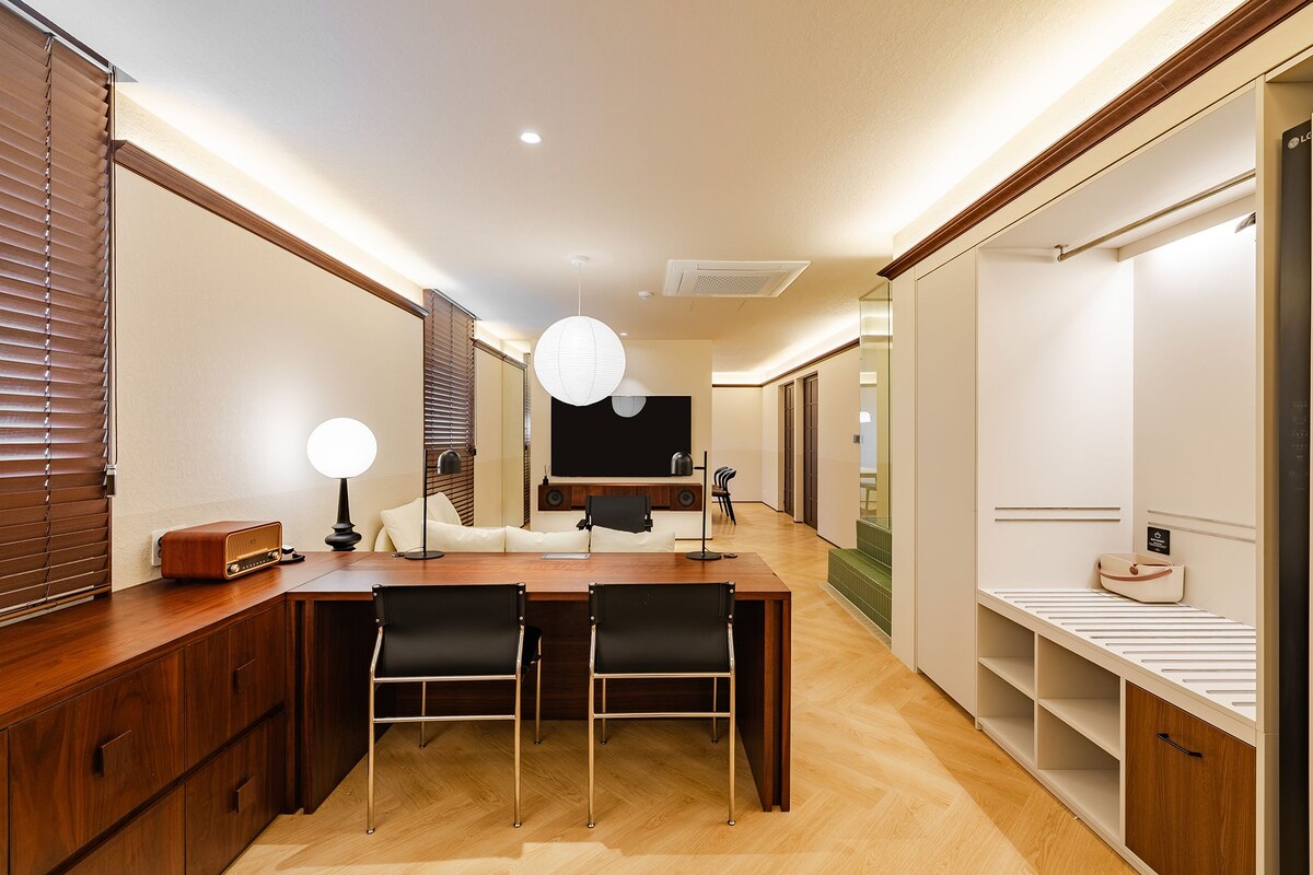 Una habitación de hotel moderna con una mesa de comedor de madera, dos sillas, un sofá, un ventanal con persianas, un televisor de pantalla plana y un armario.