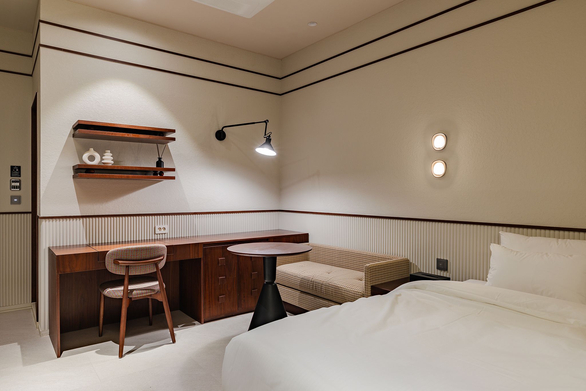 Una habitación de hotel con una cama, un escritorio con silla, una mesita, un estante en la pared y una lámpara de pared.