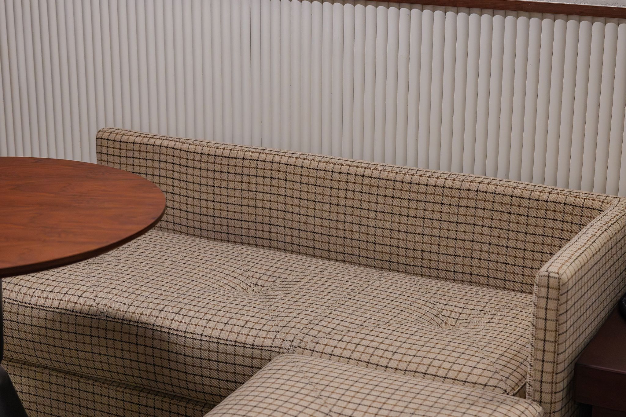 A sofa with a checkered pattern, a round wooden table, and a white vertical blinds background.