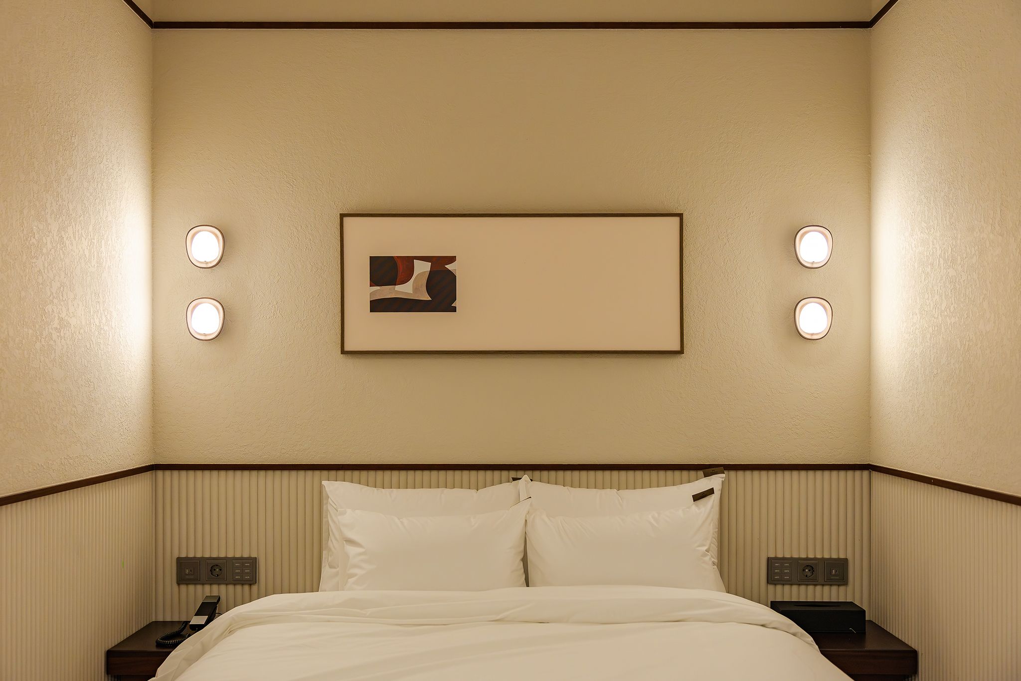 Una habitación de hotel con cama, mesitas de noche, un cuadro enmarco y lámparas fijadas a la pared.
