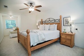 A bedroom with a wooden bed, a nightstand with a lamp, a ceiling fan, and a window with a view of trees. - Red Barn Lake House-Lake McQueeney (Seguin)