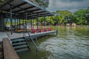 A covered dock with red chairs, a boat, and a view of a lake surrounded by trees. - Red Barn Lake House-Lake McQueeney (Seguin)