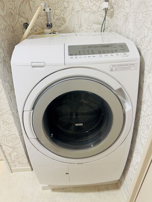 A front-loading washing machine with a control panel, set against a patterned wallpaper background. - Tokyo 3-Story Private House, Sleeps 10, Free Parking, Easy Asakusa & Airports (Katsushika City)