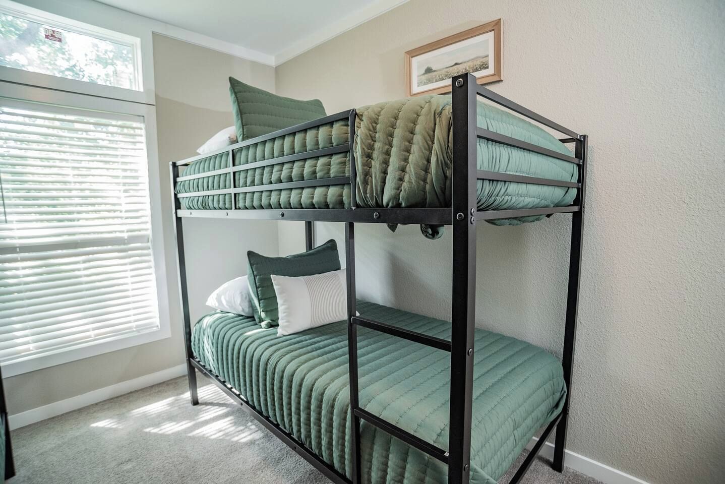 A bunk bed with green bedding and pillows, a window with blinds, and a framed picture on the wall.
