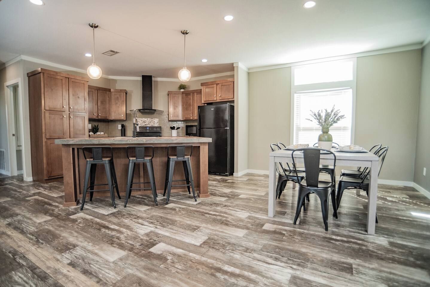 A modern kitchen with wooden cabinets, a central island with stools, a dining table with chairs, and a large window providing natural light.
