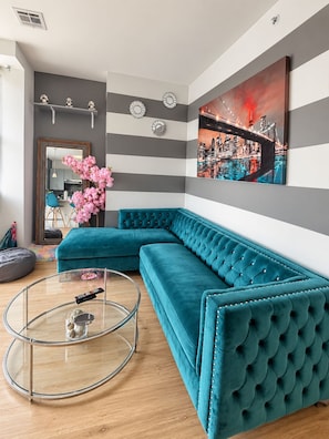 A modern living room with a teal sectional sofa, a glass coffee table, and a cityscape painting on the wall.